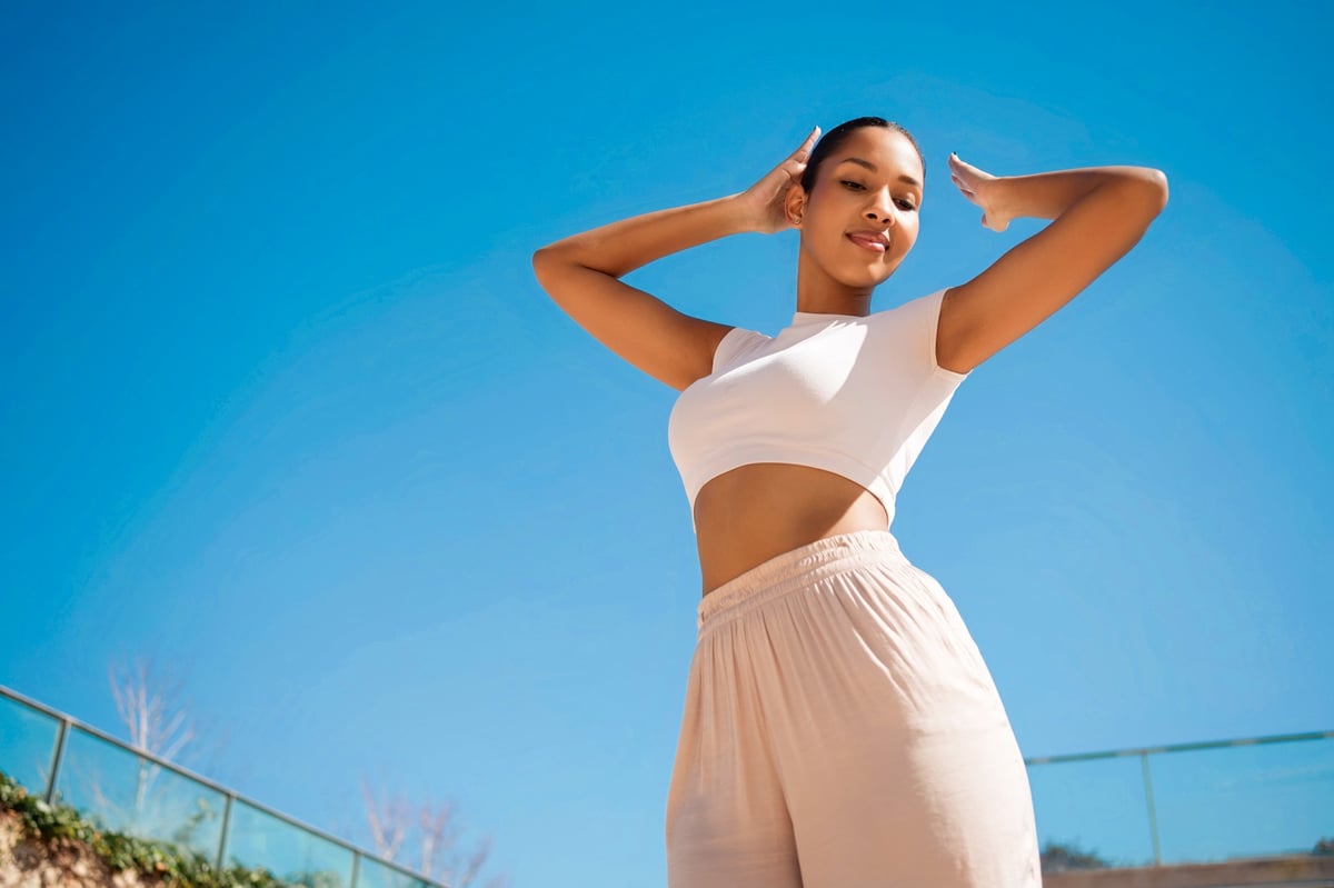 Woman stretching on a sunlit rooftop, arms raised, smiling under a clear blue sky